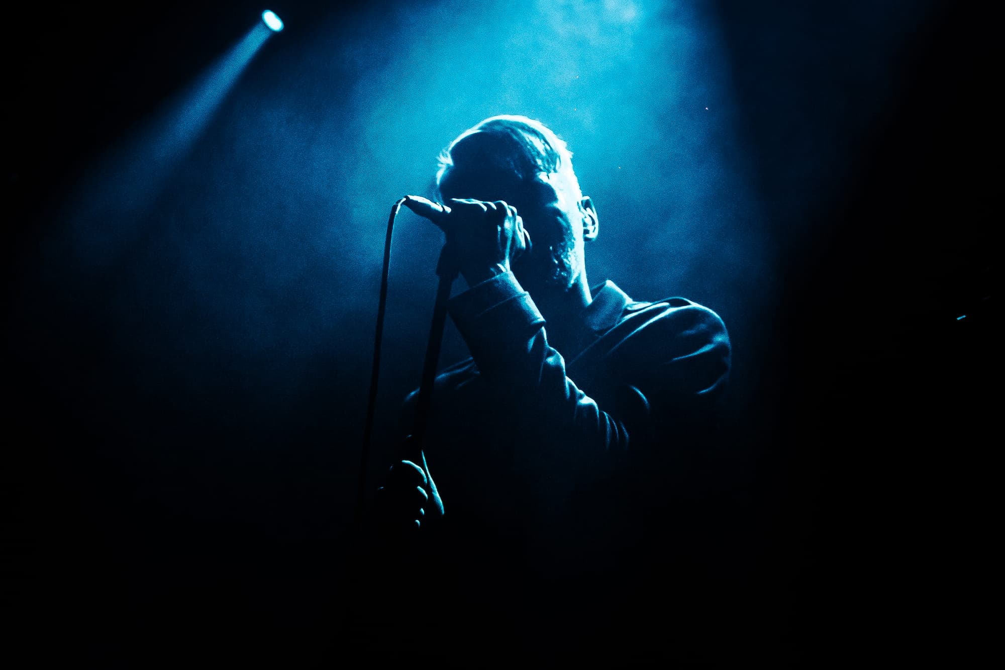 Man singing into a microphone with dark blue stage lighting behind him